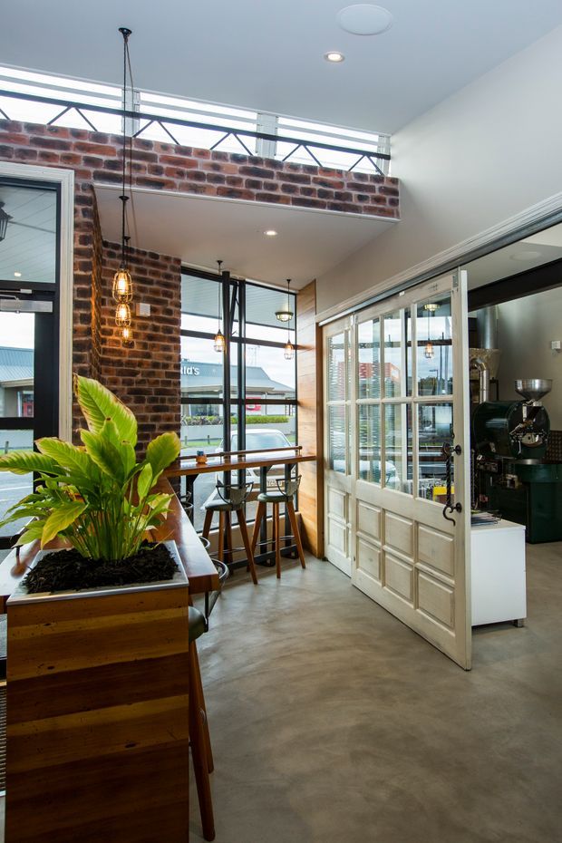 Interior view of a compact, high-ceilinged cafe or coffee shop with exposed red brick walls, polished concrete flooring, timber planter joinery, black-framed glazing, and a large glazed timber-framed partition door. Pendant lights hang above a bar-height dining counter, while a coffee machine is visible in the adjoining service area. Clerestory glazing and exposed triangular steel framing bring in abundant daylight.