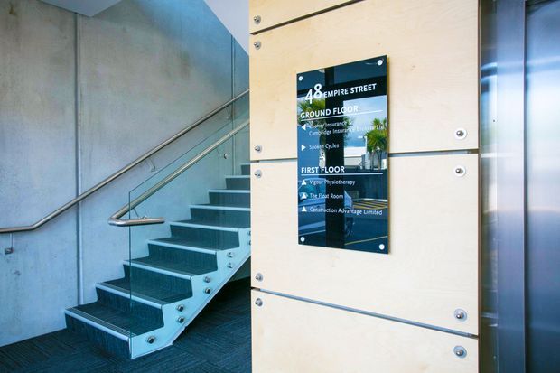 Modern commercial building entry and stairwell with a dark carpeted staircase, exposed concrete walls, stainless steel handrails, and a pale timber-panel directory sign listing businesses across the ground and first floors. A glazed lift or entrance element is visible at right.