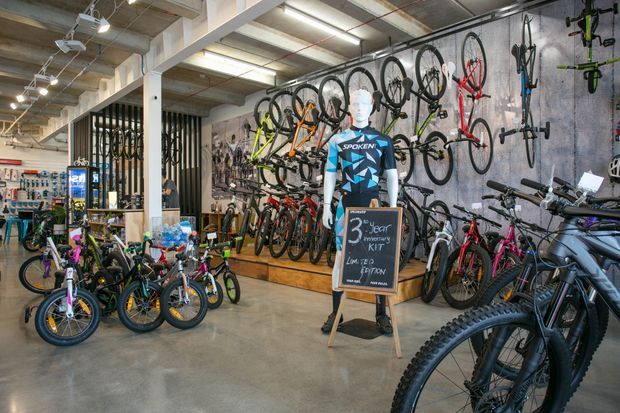Interior view of a contemporary bicycle shop with polished concrete flooring, exposed concrete and service-style ceilings, extensive wall-mounted bicycle displays, and numerous bikes arranged throughout the retail space. A mannequin and chalkboard promotional sign stand near the centre, with cycling accessories and equipment visible in the background.