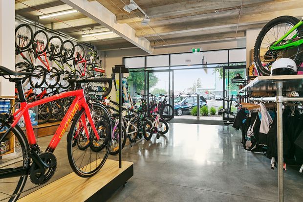 Interior view of a contemporary bicycle retail showroom, with bicycles displayed on wall racks and timber platforms, cycling apparel and accessories on shelving, polished concrete flooring, exposed timber and concrete ceiling elements, and full-height glazed frontage opening toward the street.