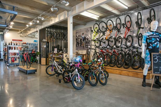 A bright, industrial-style bicycle retail showroom with polished concrete flooring, exposed concrete ceiling, structural beams, track lighting, and black metal display racks. Numerous adult and children's bicycles are displayed on wall-mounted and freestanding racks, with cycling accessories along the left wall and a mannequin wearing cycling apparel on the right.