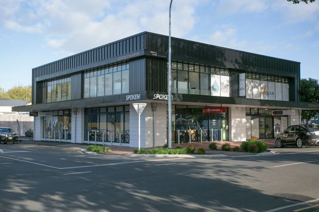 Street-level exterior photograph of a contemporary two-storey commercial building with dark metal cladding, extensive glazing, ground-floor retail tenancies, upper-level offices or studios, and a paved roadside frontage.