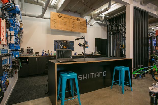 A commercial bicycle shop workshop and service counter with Shimano branding, black built-in cabinetry, exposed ceiling services and ductwork, tool storage, hanging bicycle wheels, and bright turquoise stools. The space has an industrial retail-workshop character with concrete flooring and dark metal detailing.