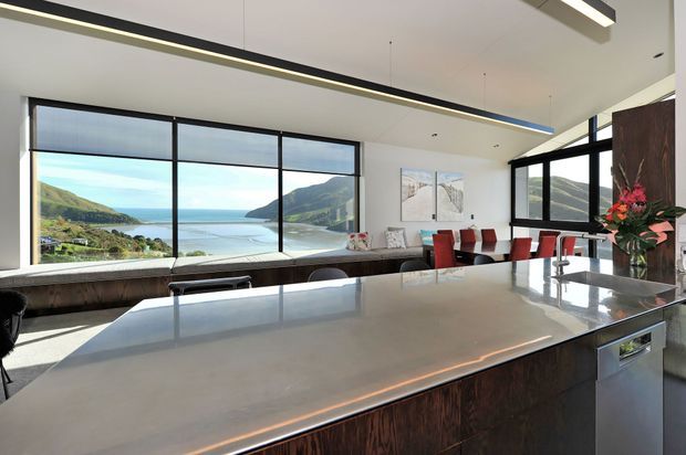 A spacious contemporary kitchen and dining area with a large stone island, dark timber cabinetry, integrated appliances, and linear pendant lighting. Floor-to-ceiling glazing provides expansive views across a coastal bay and surrounding hills. Red dining chairs, floral arrangements, and artwork add colour to the otherwise neutral, refined interior.