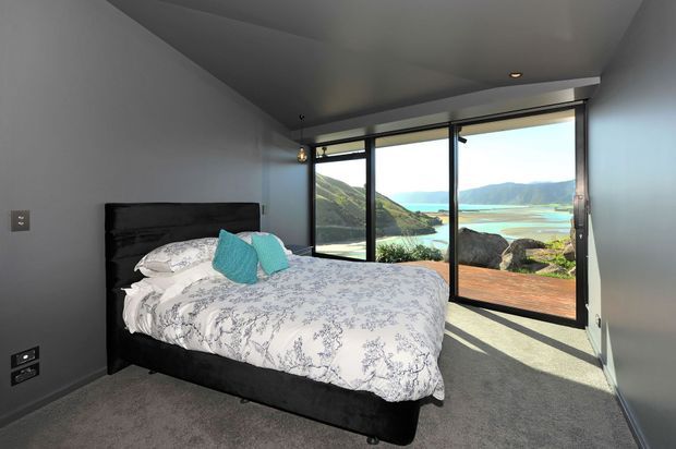 A compact modern bedroom with a black upholstered bed, patterned white bedding, and turquoise accent cushions. Dark grey walls and ceiling contrast with a soft grey carpet, while full-height black-framed glazing opens to a timber deck and a dramatic coastal landscape beyond. Minimal pendant lighting and recessed ceiling lights complete the contemporary interior.