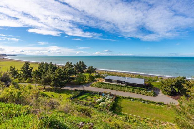A wide elevated view over a landscaped coastal property, with a low modern building set among trees and lawns beside a sandy beach and expansive turquoise ocean. The scene is dominated by natural planting, open sky, and a strong beachfront setting.