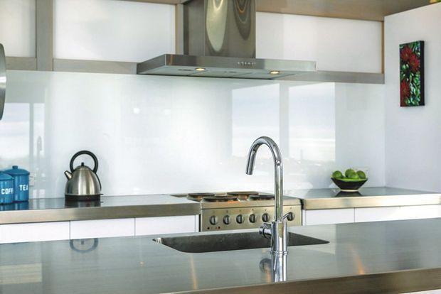 Contemporary kitchen with glossy white flat-panel cabinetry, stainless-steel benchtops and appliances, a large central island with an undermount sink and gooseneck mixer, and a stainless-steel rangehood. A reflective white splashback and restrained decor create a clean, bright look.