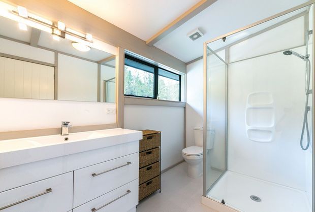 Bright contemporary bathroom with a large white vanity, integrated basin, mirrored wall, walk-in glass shower enclosure, toilet, and woven drawer storage. Light timber ceiling detailing and a black-framed window add contrast to the predominantly white interior.