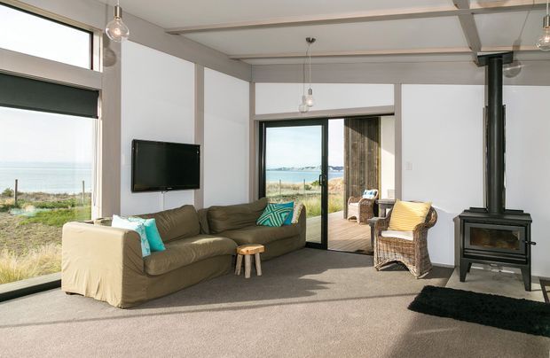 Bright coastal living room with expansive glazing overlooking a beach and ocean. The room features exposed ceiling beams, white walls, carpeted flooring, a dark freestanding wood-burning stove with flue, upholstered sofas, wicker armchairs, pendant lights, and a sliding glass door opening to a timber deck.