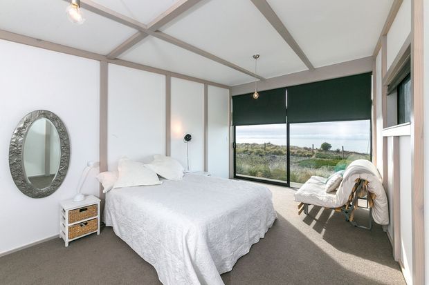 Bright contemporary bedroom with a white bed, textured white bedding, pale timber wall and ceiling framing, grey carpet, and expansive black-framed glazing overlooking the landscape. A woven bedside table, sculptural round mirror, lounge chair, and simple pendant lighting create a relaxed, airy interior.