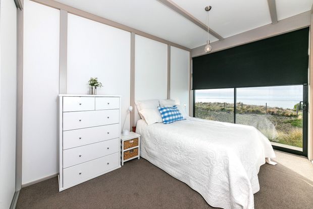 Bright, compact bedroom with a single bed dressed in white bedding, a blue checked accent cushion, white chest of drawers, bedside table, and large floor-to-ceiling windows overlooking a coastal landscape. The room features white panelled walls with pale timber trim, exposed ceiling beams, dark carpet, and a dark roller blind.