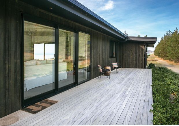 Exterior view of a low-profile contemporary home with dark vertical timber cladding, expansive black-framed sliding glass doors, and a broad pale timber deck. Outdoor chairs sit on the deck beside landscaped planting, while a bedroom and ocean or bright horizon are visible through the glazing and reflections.