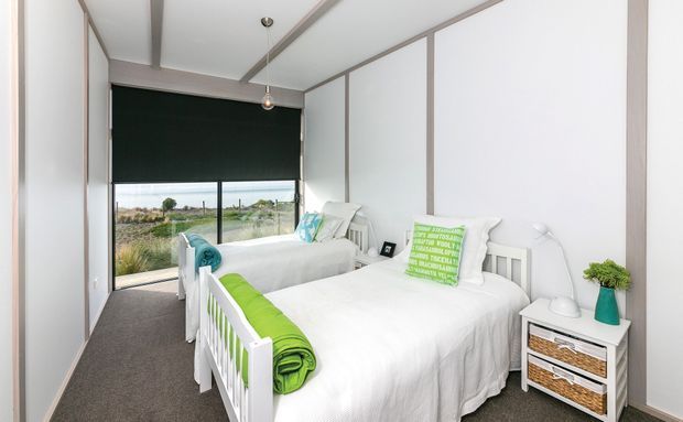 Bright shared bedroom with two white beds, white panelled walls and ceiling, grey carpet, and full-height glazing overlooking an outdoor landscape. Green and turquoise accents appear in the bedding, cushions and accessories, with compact white bedside storage and simple pendant lighting.