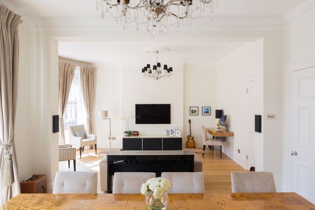 Bright, refined open-plan living and dining interior with white walls, ornate ceiling cornices, tall curtained windows, decorative chandeliers, a black wall-mounted TV and media console, piano, desk, and a large timber dining table with upholstered chairs. The room has a classic period character balanced with contemporary furnishings.