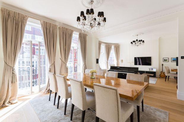 Bright, elegant open-plan dining and living space with a long polished timber dining table surrounded by upholstered chairs, tall curtained windows opening to a balcony, light timber flooring, ornate ceiling mouldings, and crystal chandeliers. A minimalist TV console and seating area are visible beyond the dining zone.