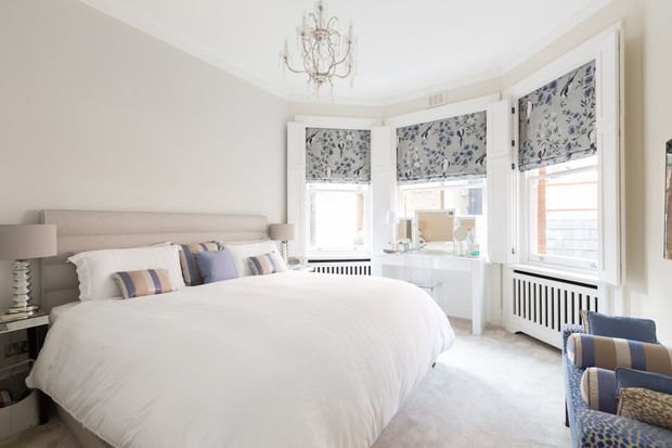 Bright, refined bedroom with a large upholstered bed dressed in white bedding, pale neutral walls, and a curved bay window with patterned Roman blinds. A delicate chandelier, bedside lamps, compact dressing desk, radiator covers, and two blue patterned armchairs complete the elegant interior.