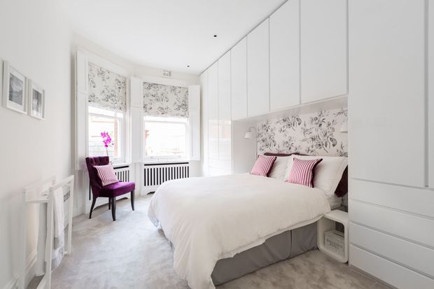 Bright, refined bedroom with a large upholstered bed, white bedding, pink striped accent cushions, floral patterned roller blinds and a matching floral feature wall behind the bed. Floor-to-ceiling white built-in wardrobes line the right side, while a purple upholstered chair sits beside tall windows. The room has a soft, elegant, predominantly white and blush palette.