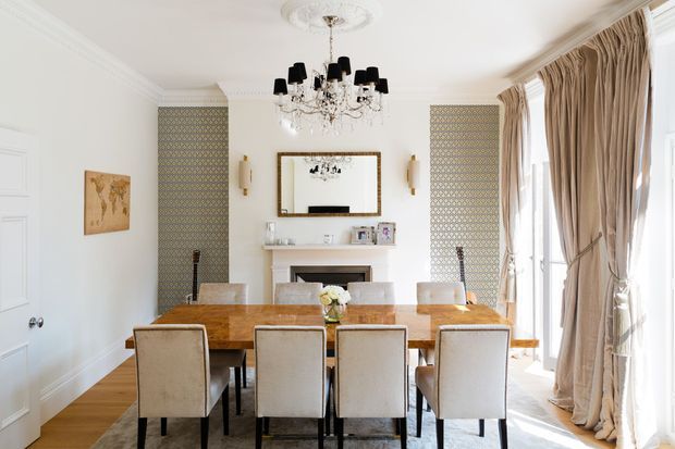 Bright formal dining room with a large polished timber dining table, upholstered dining chairs, a decorative fireplace and mirror, patterned feature wall panels, chandelier, wall sconces, and full-height draped windows. The room has refined traditional detailing with a warm, elegant palette.