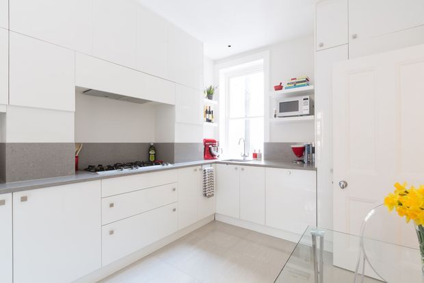 Bright, compact contemporary white kitchen with extensive handleless-style cabinetry, a grey stone-look benchtop and splashback, integrated appliances, gas cooktop, and a central window above the sink. A glass dining table and transparent chair are partially visible in the foreground.