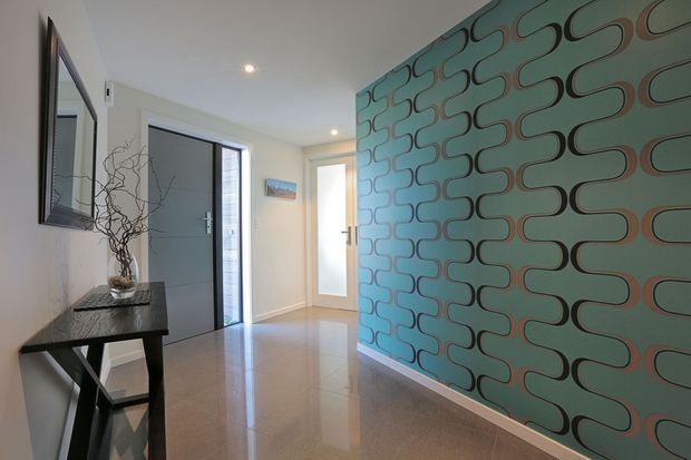 A bright contemporary entry hall with a polished tiled floor, grey front door and adjacent glazed side panel, a console table with decorative branches and mirror, and a bold teal geometric feature wall with looping black and terracotta pattern. Recessed ceiling lights and a secondary interior door complete the space.