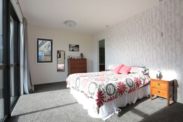 A bright bedroom with a large floral-patterned bedspread, white bed skirt, grey carpet, and patterned feature wallpaper. A timber dresser and bedside table provide warm wood accents, while full-height glazed doors with curtains bring in abundant natural light. Framed artwork and a mirror complete the room.