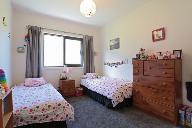 A cheerful shared children's bedroom with two single beds dressed in colourful patterned bedding, a central bedside table, grey carpet, curtained window, and a large wooden chest of drawers. Children's artwork, toys, and personal items add a playful character.
