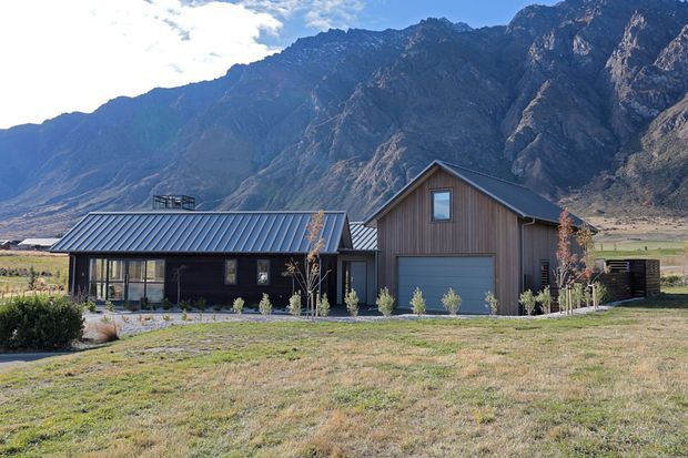A contemporary mountain home with dark vertical timber cladding, a low-pitched metal-roofed single-storey wing, and a taller gabled garage or living volume. The house sits in an open grassy valley backed by dramatic rocky mountains. Large glazed openings, a covered entry connection, and a simple landscaped frontage are visible.