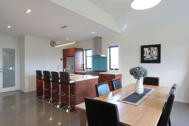 Bright open-plan kitchen and dining area with polished flooring, dark timber kitchen cabinetry, a central island with four bar stools, stainless-steel appliances, and a light timber dining table with black upholstered chairs. White walls, recessed lighting, pendant lighting, and contemporary artwork create a clean modern interior.