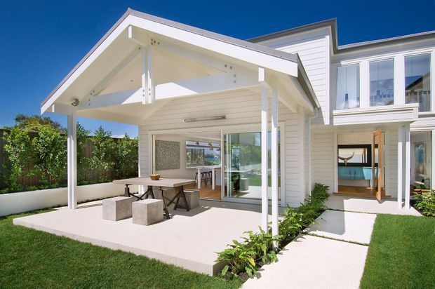 Bright exterior view of a contemporary white weatherboard home with a prominent covered outdoor entertaining terrace, exposed gable roof structure, large glazed doors, and a landscaped lawn. The terrace includes an outdoor dining setting and built-in concrete seating, while the interior living spaces are visible through the glazing.