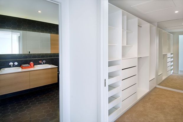 A bright dressing area with extensive white built-in shelving, drawers, hanging compartments and mirrored wardrobe doors, opening directly onto a contemporary bathroom. The bathroom features dark hexagonal wall and floor tiles, a timber-veneer vanity with twin white basins, wall-mounted chrome tapware and a large mirror. Neutral carpet and white joinery create a clean, functional interior.