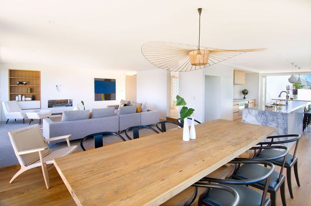 Bright open-plan contemporary interior featuring a large timber dining table with black-framed chairs in the foreground, a grey sectional sofa and occasional seating in the living area, and a marble-look kitchen island beyond. White walls and ceilings, pale timber flooring, built-in shelving, indoor greenery, and a sculptural oversized pendant fan define the airy space.