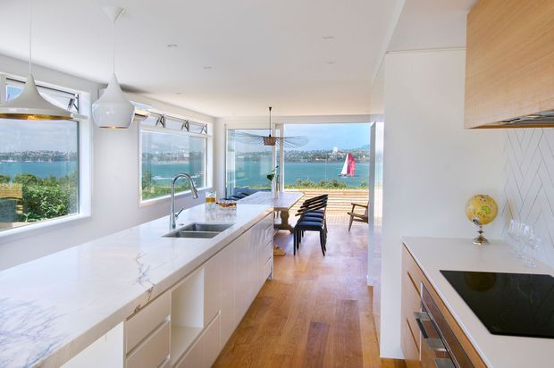 Bright contemporary open-plan kitchen with a long white marble-look island, integrated sink, white cabinetry, pendant lights, timber flooring, and a white herringbone splashback. Full-height glazing and an open connection to the dining area provide expansive waterfront views, with a sailboat visible outside.