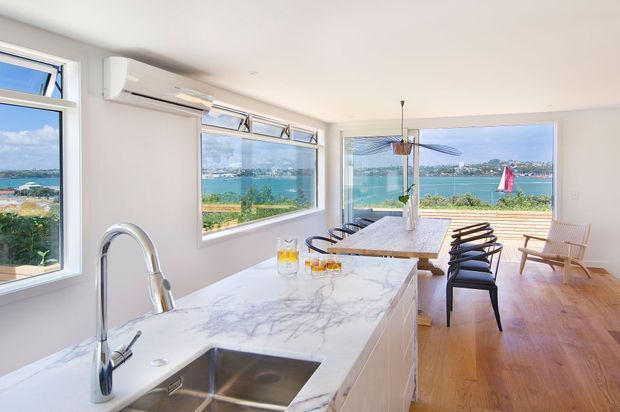 Bright open-plan kitchen and dining area with expansive waterfront views through large windows and sliding glass doors. The room features a white marble-look kitchen island with an integrated stainless-steel sink and curved chrome tap, white walls and ceiling, warm timber flooring, a timber dining table, and dark dining chairs. An outdoor deck and water are visible beyond the glazing.
