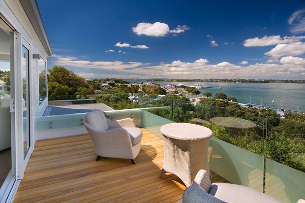 Elevated outdoor terrace with timber decking, frameless glass balustrading, and light woven outdoor seating overlooking a broad waterfront inlet and coastal suburban landscape. The open, sunny setting features a relaxed seating arrangement and expansive water views.
