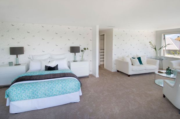 Bright, spacious bedroom with a white upholstered bed, turquoise patterned bedding, matching bedside tables and lamps, delicate bird-patterned wallpaper, and a small white sofa seating area by the window. Neutral carpet and white furnishings create a light, airy coastal feel.