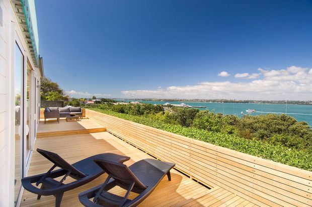 A spacious timber deck extends along the side of a light-coloured coastal residence, with built-in timber screening and steps between levels. Dark reclining lounge chairs sit in the foreground, while a turquoise bay, boats, and distant shoreline are visible beyond dense greenery under a bright blue sky.