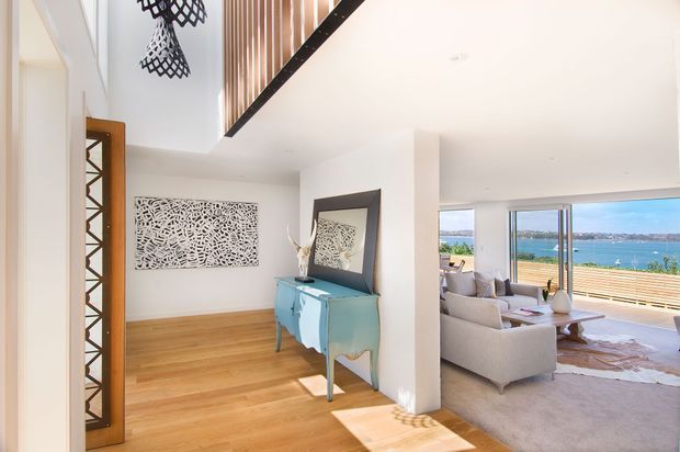 Bright contemporary open-plan interior with a timber-floored entry or circulation area leading into a living room. White walls and ceiling, a sculptural patterned wall artwork, a turquoise console, vertical timber screening, and expansive glazing overlooking blue water create an airy coastal feel.