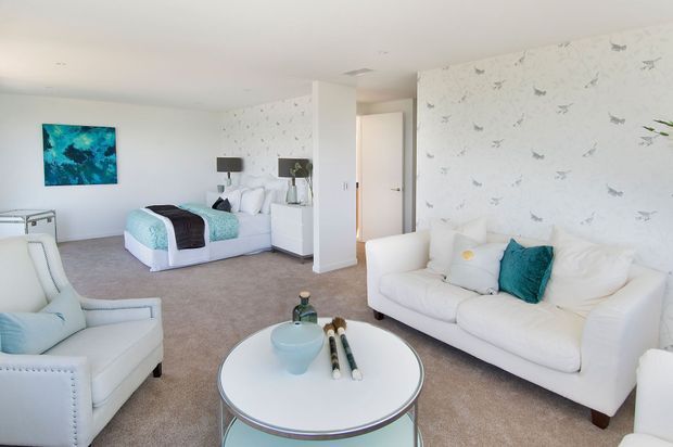 Bright, airy bedroom suite with a white upholstered bed, matching white seating, pale carpet, and patterned wallpaper featuring delicate grey motifs. Turquoise and blue accents appear in the bedding, cushions, artwork, and decorative objects. A sitting area and coffee table occupy the foreground, while built-in cabinetry and a doorway are visible behind the bed.