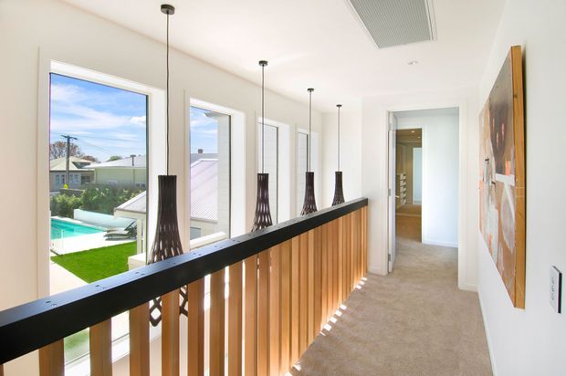 Bright contemporary upstairs hallway or landing with a long timber-slat balustrade, black metal handrail, and a row of sculptural pendant lights. Full-height windows overlook a backyard and swimming pool, while a carpeted corridor leads toward an interior doorway. White walls and ceiling create an airy, modern setting, with a large artwork mounted on the side wall.