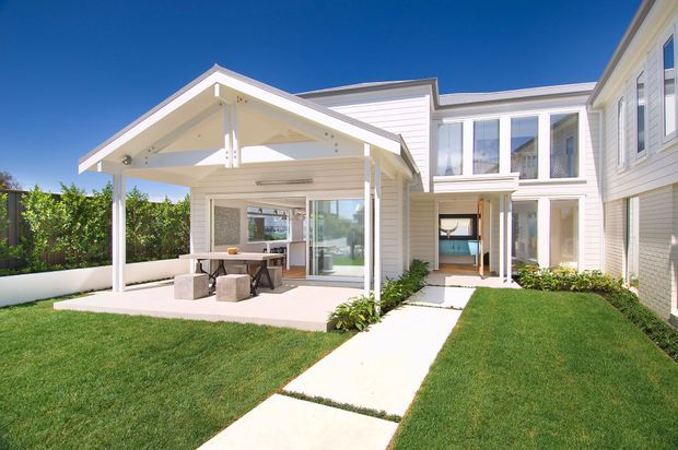 Bright exterior view of a white, multi-wing residential home with horizontal weatherboard cladding, gabled roof forms, large glazed doors and windows, and a covered outdoor dining or entertaining pavilion opening onto a manicured lawn. A concrete path leads through the landscaped courtyard toward the house.