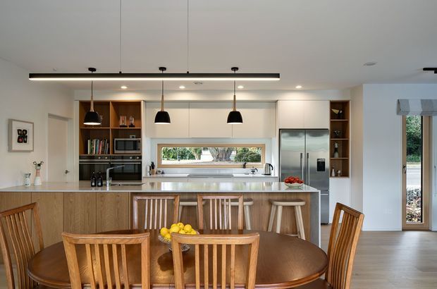 Bright open-plan kitchen and dining area with a long island, integrated appliances, pale cabinetry, timber drawer fronts, pendant lighting, and a round timber dining table with matching chairs. A wide horizontal window above the benchtop and a glazed exterior door bring in natural light. The space has a warm, clean, contemporary feel with natural timber accents.