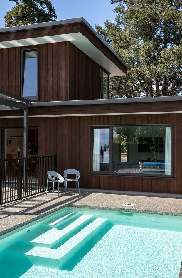 Exterior view of a contemporary multi-level home beside a swimming pool. The facade is clad in dark vertical timber boards, with broad flat roof overhangs, large dark-framed windows, and a covered outdoor terrace. Pool steps and turquoise water dominate the foreground, while mature trees surround the property.