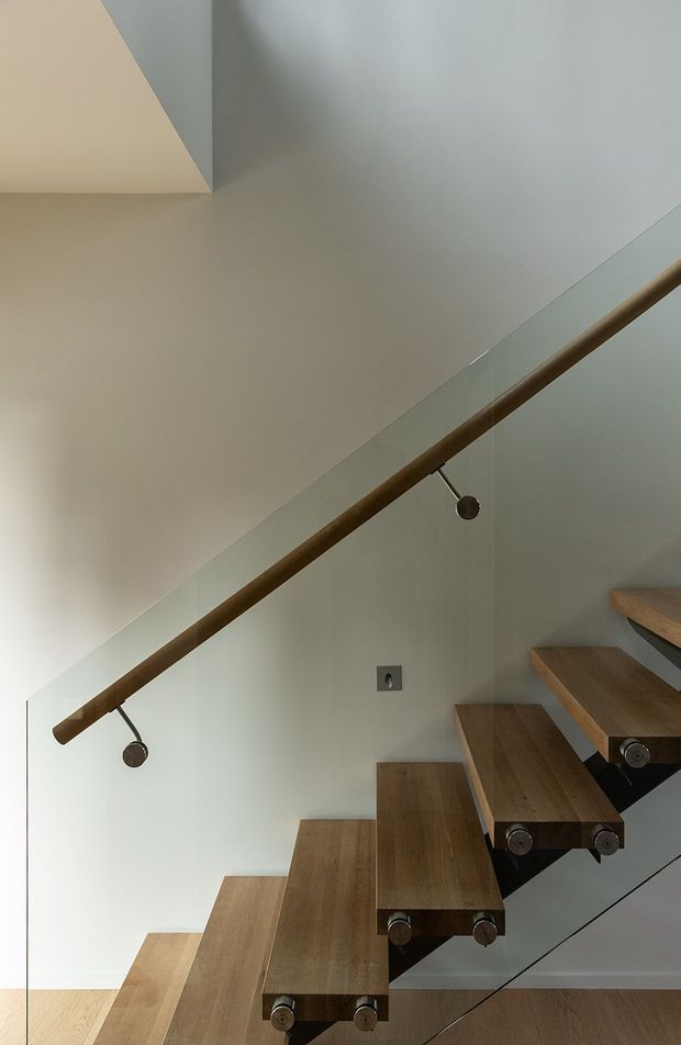 Close-up architectural photograph of a contemporary floating staircase. Thick timber treads are cantilevered from the wall with visible dark metal supports and circular fittings. A clear glass balustrade follows the stair edge, topped by a warm timber handrail with minimalist metal detailing. Pale grey walls and light timber flooring create a clean, restrained interior.