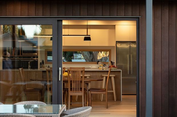 View through a large dark-framed sliding glass door into a warm, compact kitchen and dining area. The interior features timber cabinetry and wall finishes, a long island or breakfast bench with timber stools, a dining table with wooden chairs, integrated stainless-steel refrigeration, a black pendant light, and a horizontal garden-facing window. Dark vertical timber cladding surrounds the opening, while outdoor furniture is visible in the foreground.