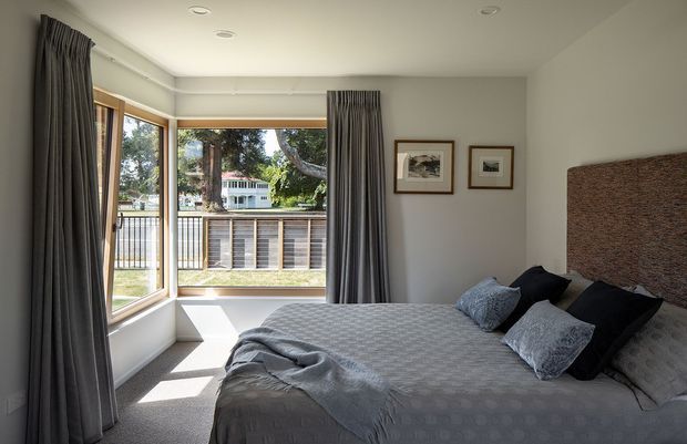 A bright, contemporary bedroom with a large corner window overlooking greenery and neighbouring homes. The room features a grey upholstered bed with patterned bedding, dark charcoal curtains, pale grey walls, carpeted flooring, recessed ceiling lights, and a textured woven-style headboard. Framed artwork adds a restrained decorative touch.