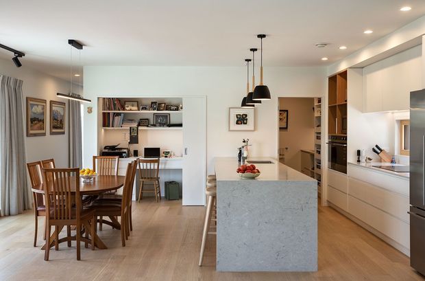 Open-plan kitchen and dining area with a central stone island, integrated white cabinetry, timber flooring, pendant lights, and a timber dining table. Built-in shelving with books and framed photographs is visible behind the dining area, with a hallway and additional kitchen joinery beyond.