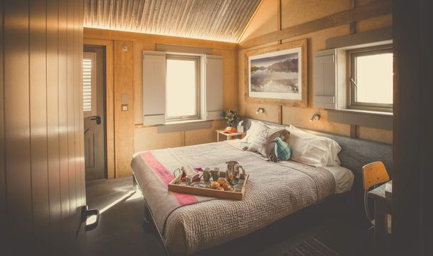 A warm, compact bedroom with a neatly made bed, timber-lined walls and ceiling, built-in wardrobes, a framed landscape print, and windows with shutters. The room has a cosy cabin-like atmosphere with muted neutral bedding, warm wood tones, and a small yellow chair at a desk or side table.