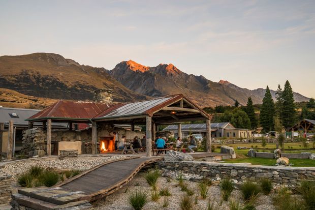 A rustic mountain property with a covered timber-framed outdoor shelter, corrugated metal roof, stone fireplace and seating area. A timber walkway crosses a landscaped gravel garden with low stone retaining walls, ornamental grasses and raised garden structures, backed by dramatic alpine peaks.