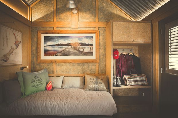 A compact, warmly lit bedroom with a built-in daybed beneath framed artwork. The room features rustic timber framing, textured warm-toned wall panels, a corrugated metal sloped ceiling, integrated shelving and hanging storage, plantation shutters, and layered bedding with casual cushions. The overall atmosphere is cozy, earthy, and cabin-like.