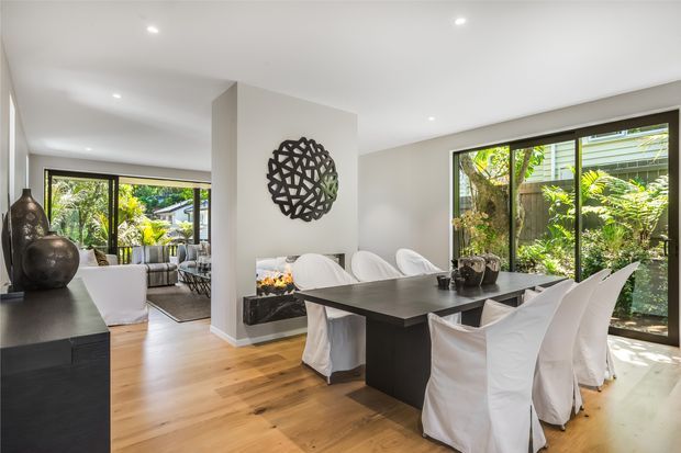 Contemporary open-plan dining and living area with a large dark rectangular dining table, white slipcovered dining chairs, light timber flooring, and white walls and ceiling. Floor-to-ceiling black-framed glazing opens views to lush greenery, while a sculptural dark wall feature and dark sideboard add contrast.